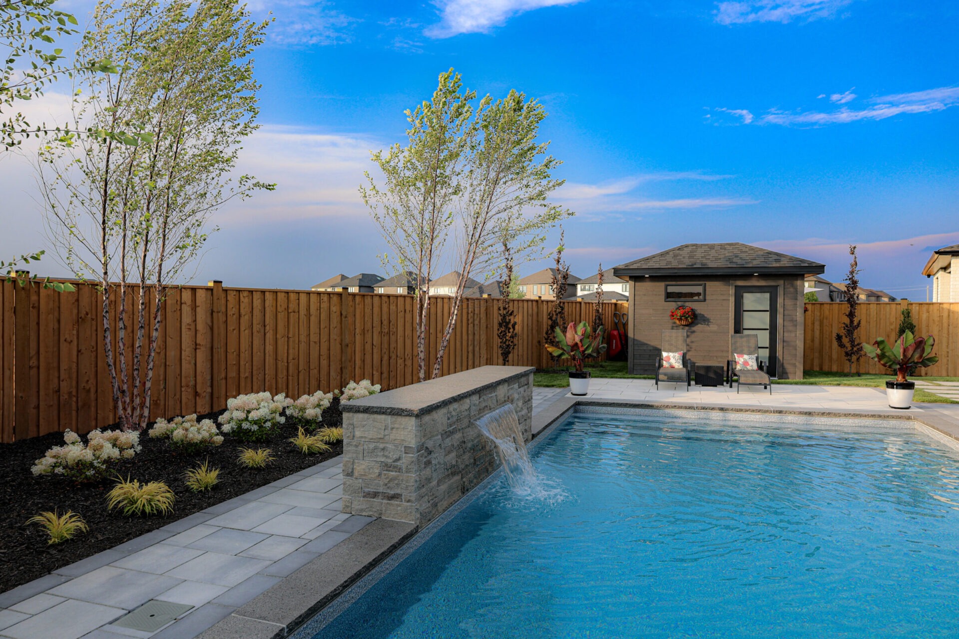 A backyard features a pool with a waterfall, two chairs, potted plants, and a small building, all under a clear blue sky.