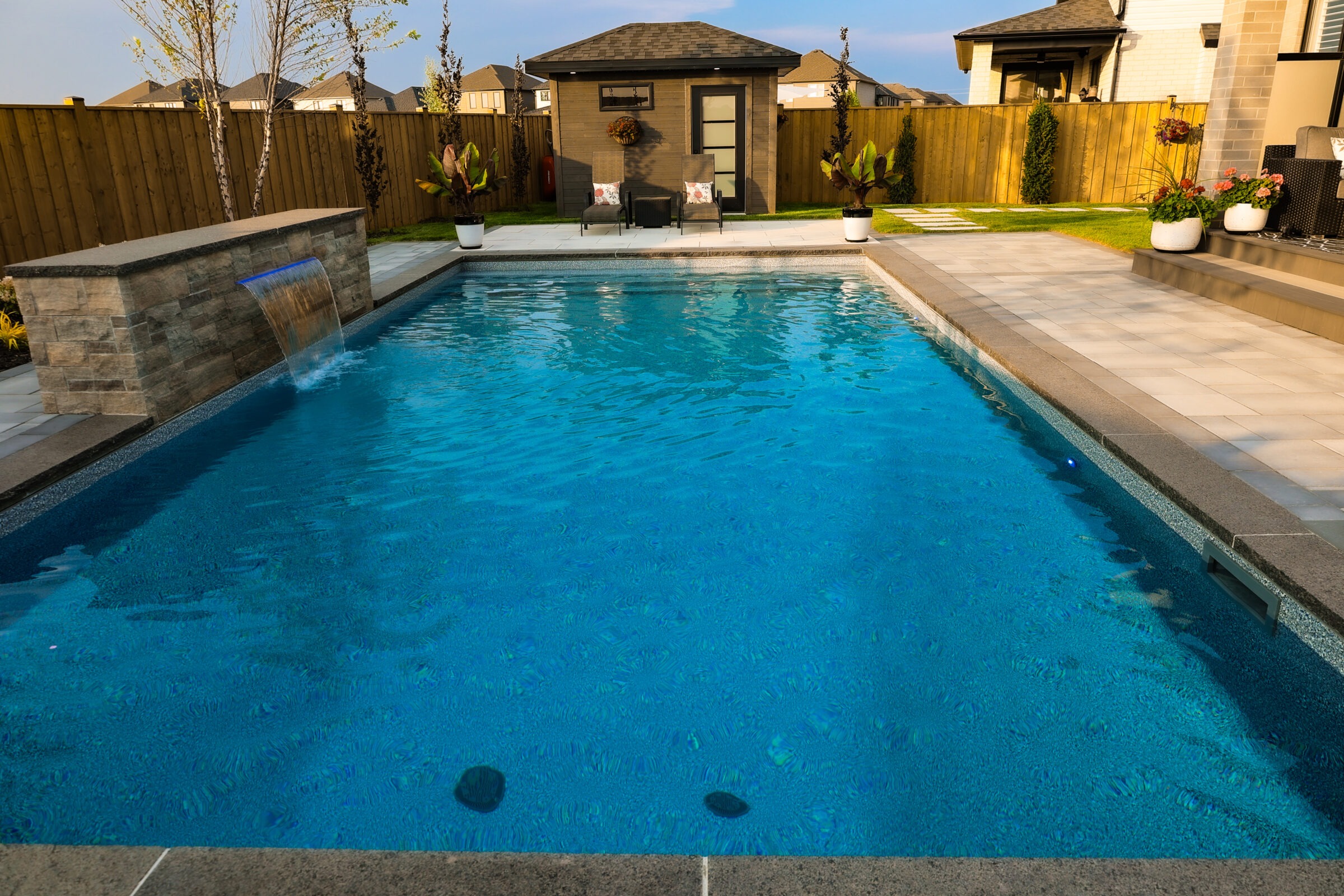 Rectangular backyard pool with waterfall feature, surrounded by modern patio and chairs. Fenced with greenery, small building present. Peaceful, suburban setting.