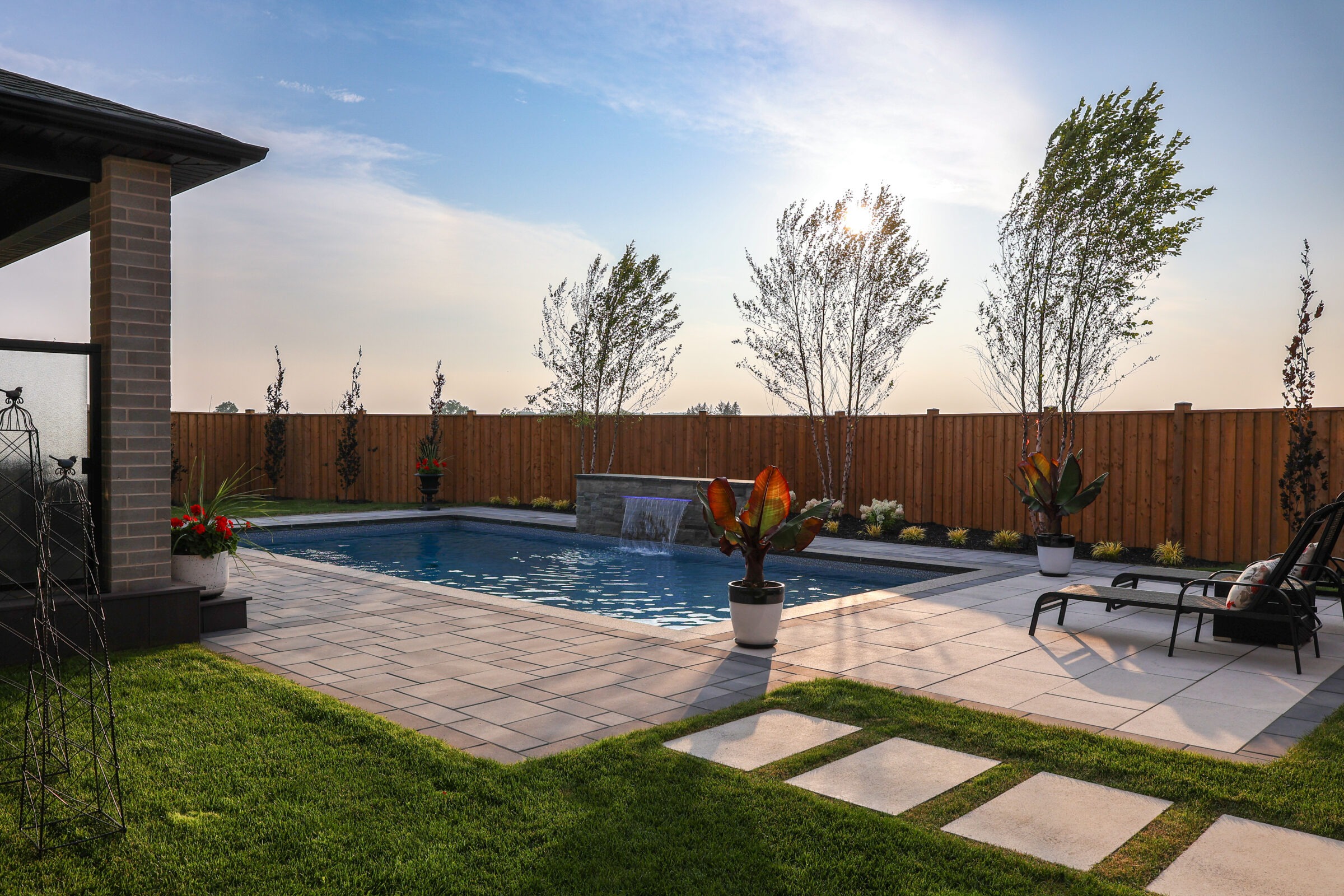 A backyard with a swimming pool, tiled patio, potted plants, and lounge chairs, enclosed by a wooden fence under a clear sky.