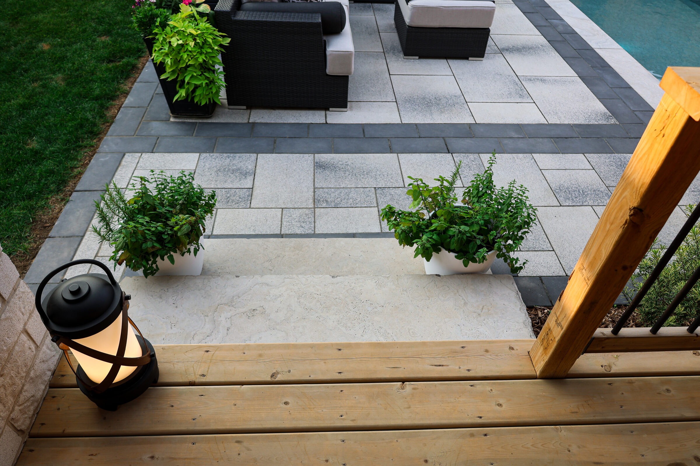 Modern patio with wooden steps, lantern, potted plants, and comfortable seating near a pool. Geometric tile design adds a stylish touch.