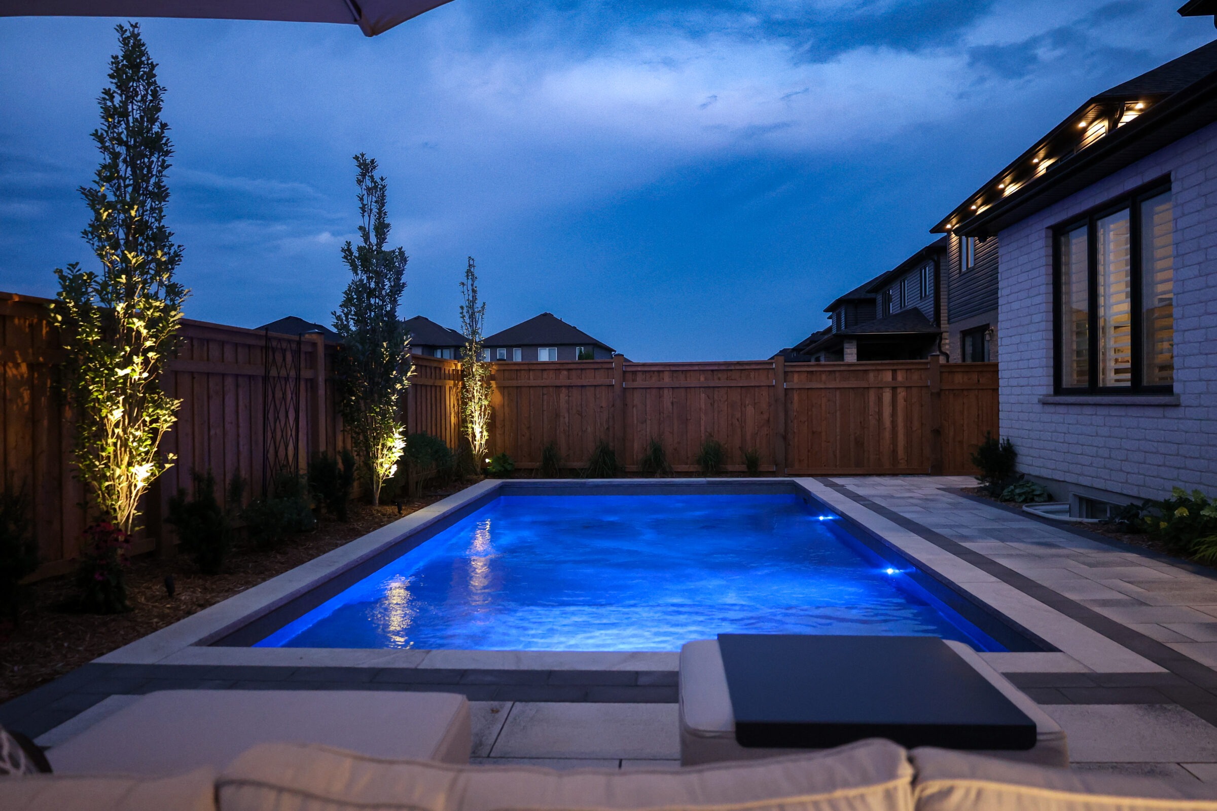 A serene backyard scene at dusk features an illuminated swimming pool, surrounded by wooden fencing and neatly landscaped trees under a deep blue sky.