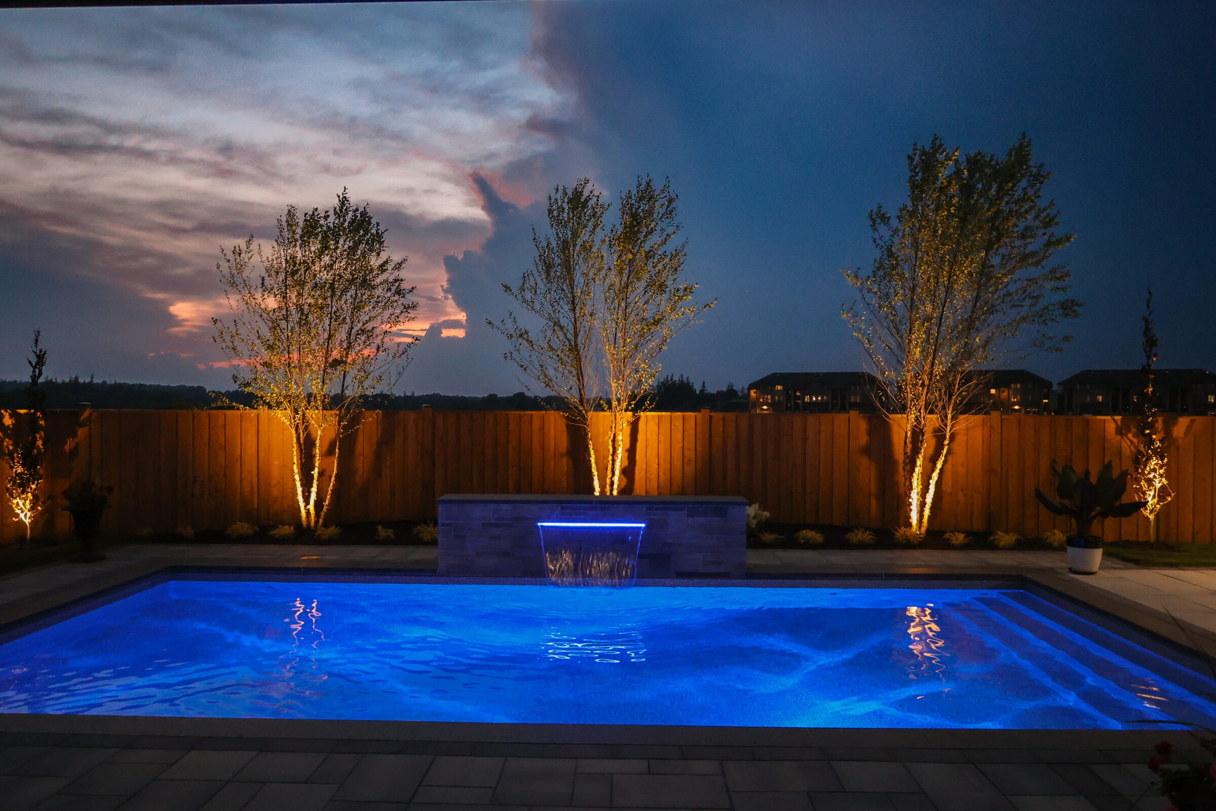 A backyard featuring a lit rectangular swimming pool at dusk, surrounded by trees and a wooden fence, creating a serene evening atmosphere.