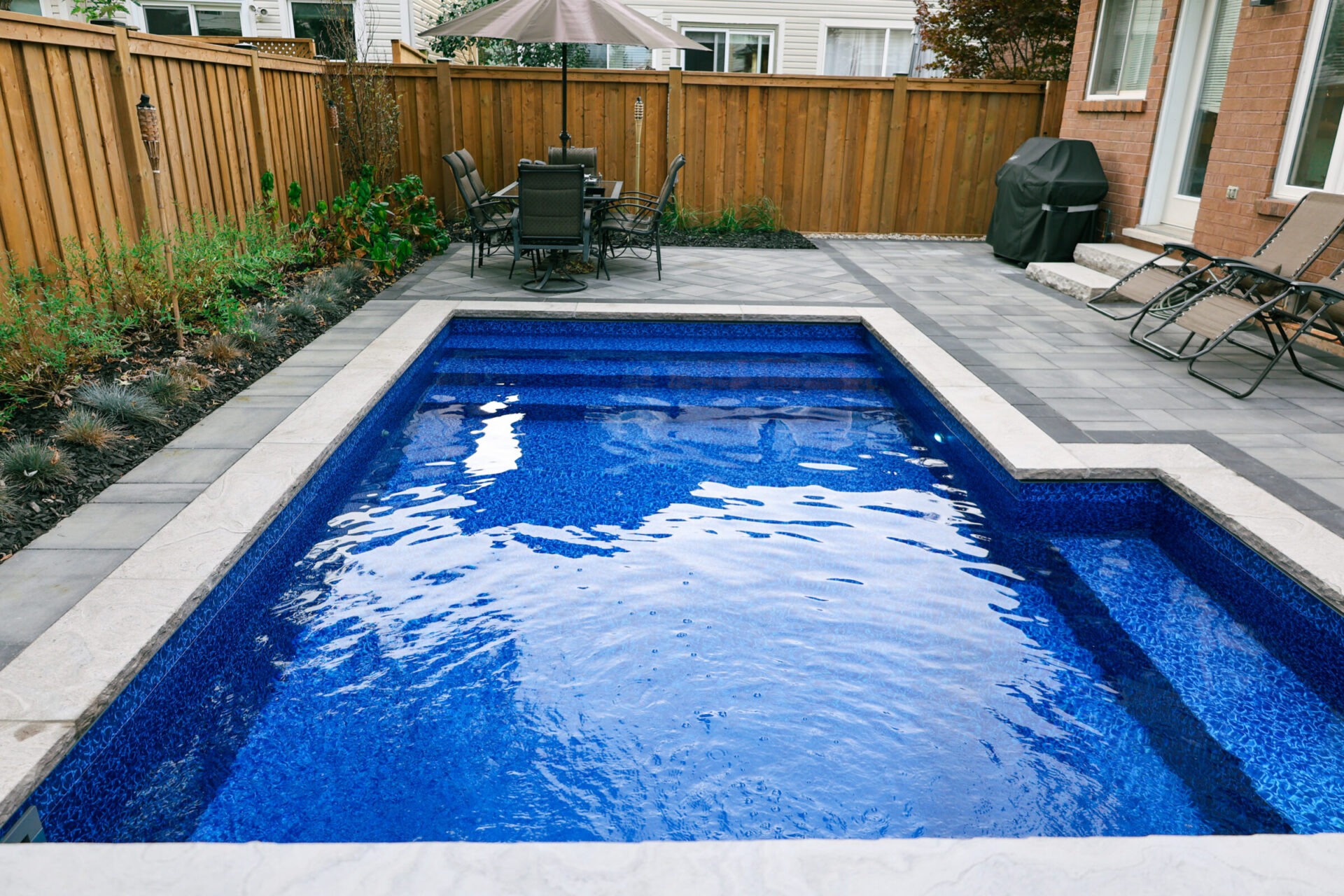 Backyard features a small, rectangular pool with blue tiles, surrounded by a patio area, dining set, lounge chairs, and a barbecue grill.