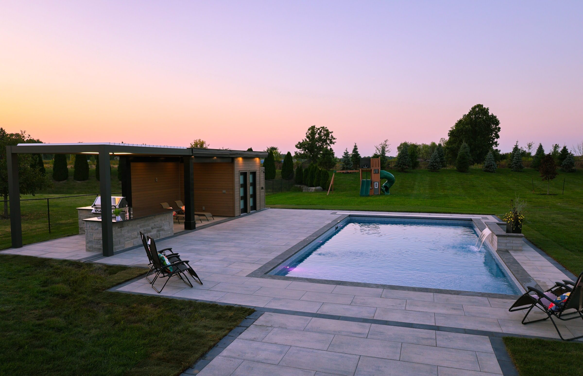 A modern backyard features a swimming pool, pergola, and playground, set against a serene sunset sky with spacious green lawn.