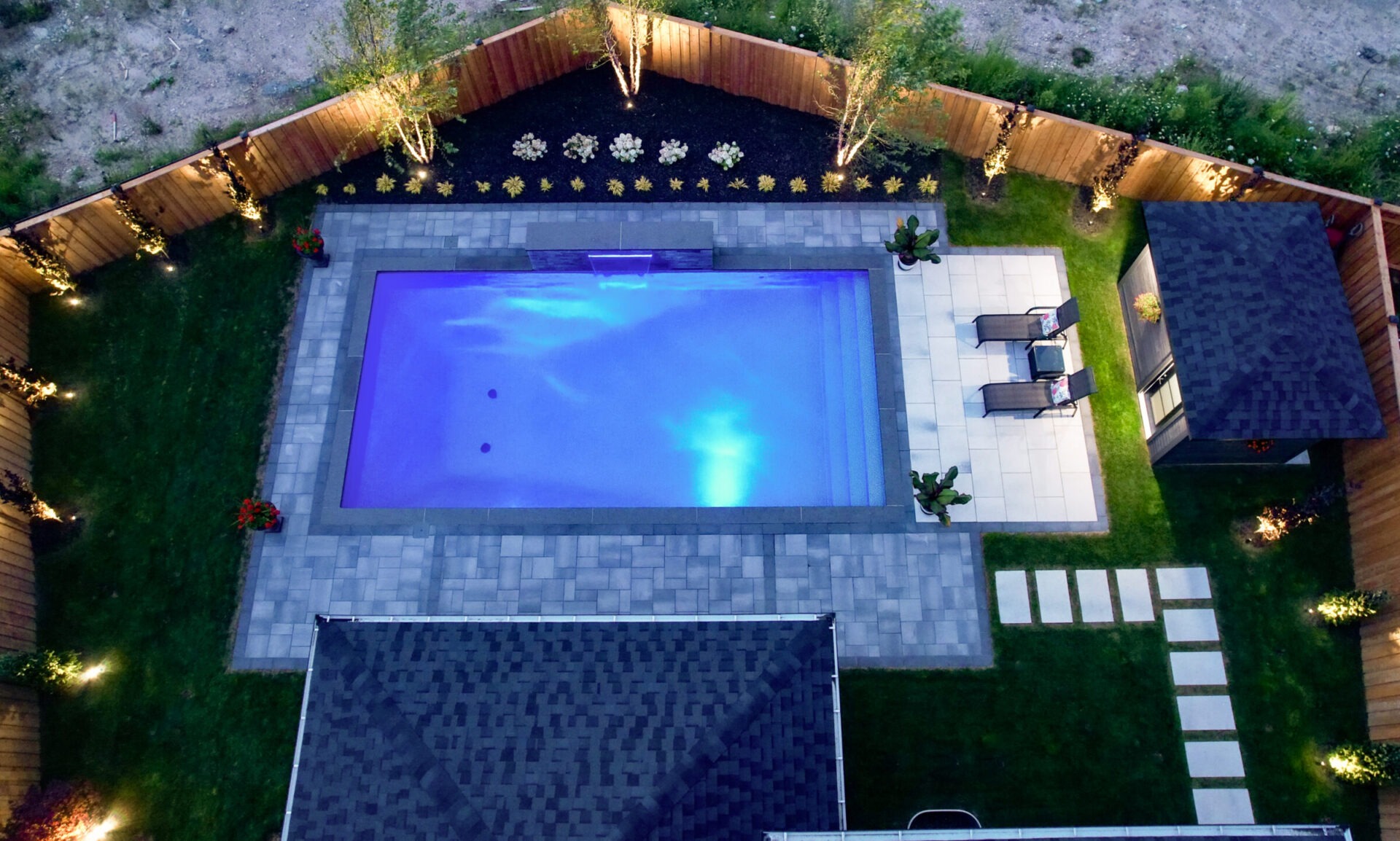 Aerial view of a lit backyard pool, surrounded by patio, lounge chairs, small shed, and landscaped garden with decorative lighting at night.