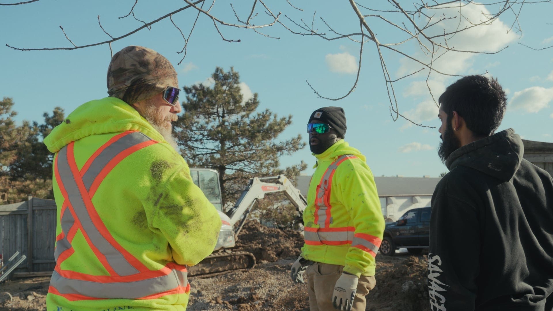 Three persons in safety gear converse at a construction site with a small excavator, surrounded by trees and a clear sky.