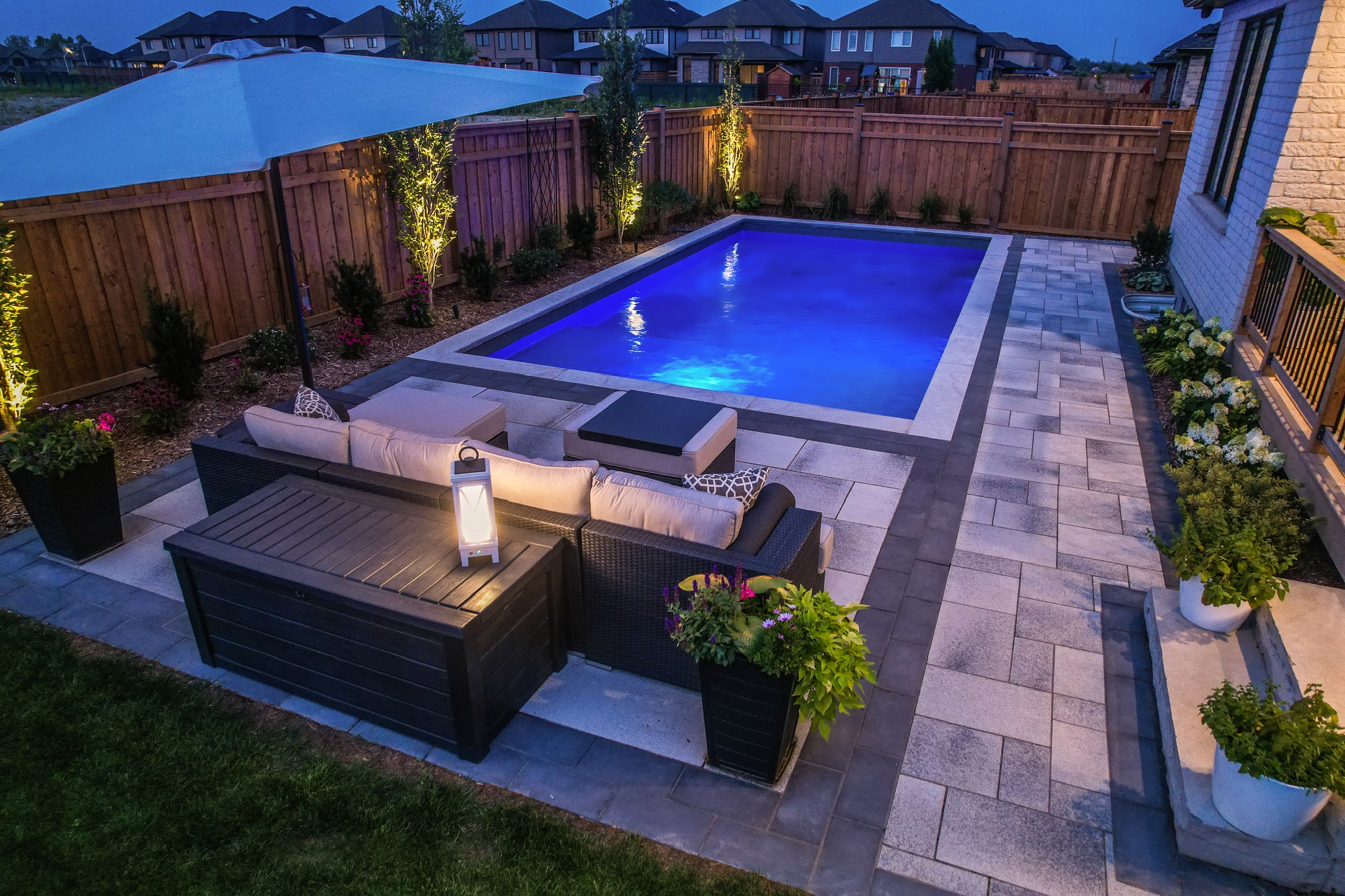 A cozy backyard features a lit swimming pool, cushioned seating, a patio umbrella, and potted plants, surrounded by a wooden fence.
