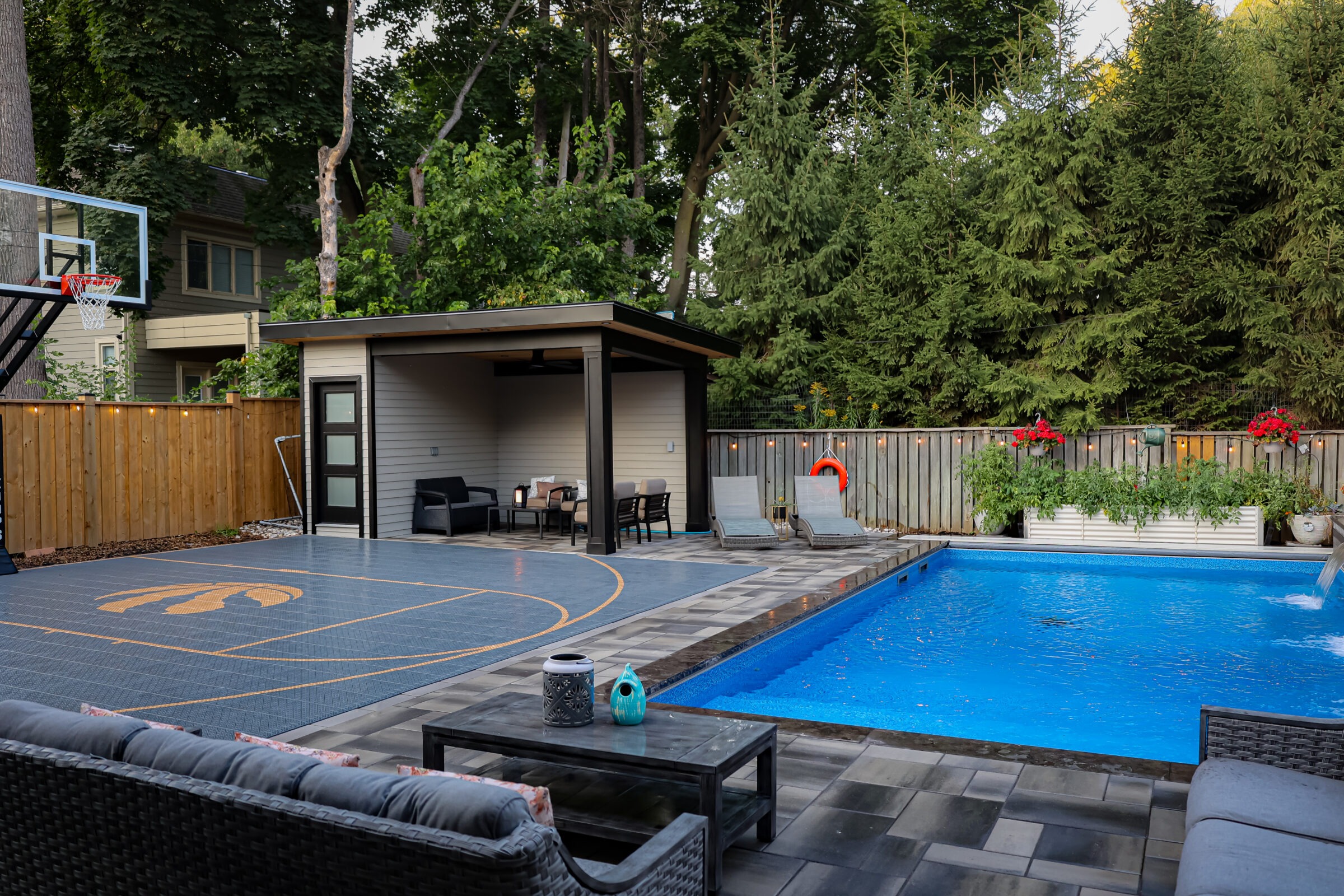 Backyard features a basketball court, swimming pool, and furnished patio. Surrounded by trees and a wooden fence, creating a cozy outdoor space.