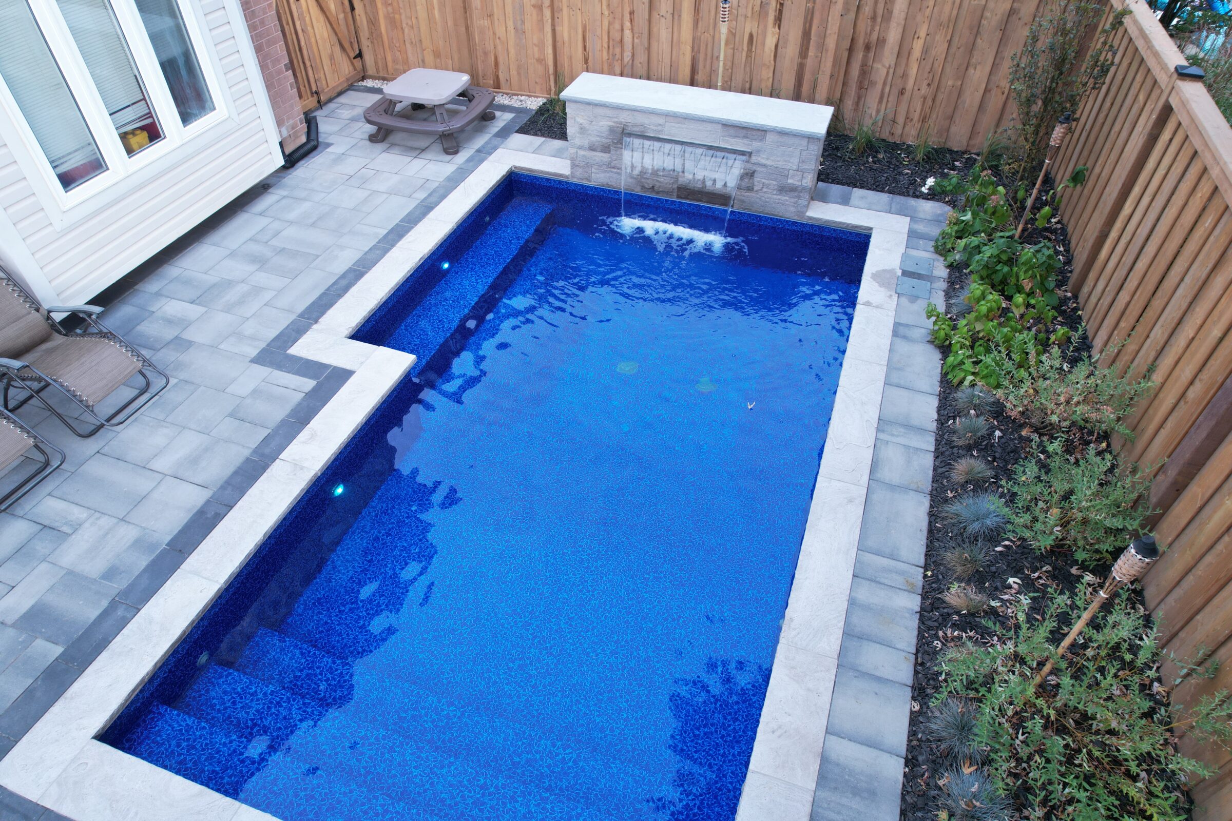 A small, rectangular backyard pool with surrounding patio, wooden fence, plants, and seating area. Clean, modern design with a built-in waterfall.