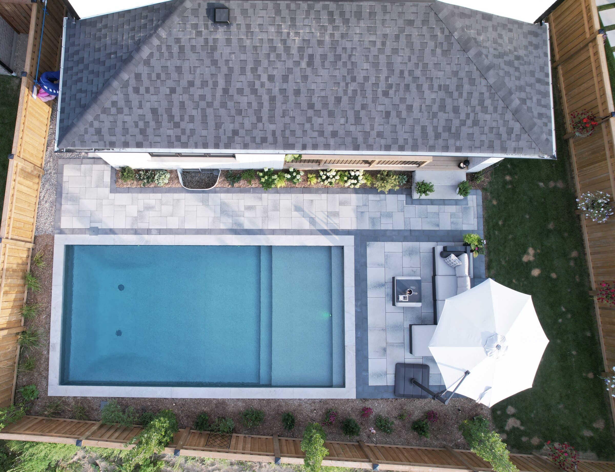 Aerial view of backyard with a rectangular swimming pool, patio, umbrella, seating area, and greenery. A person stands near the wall.