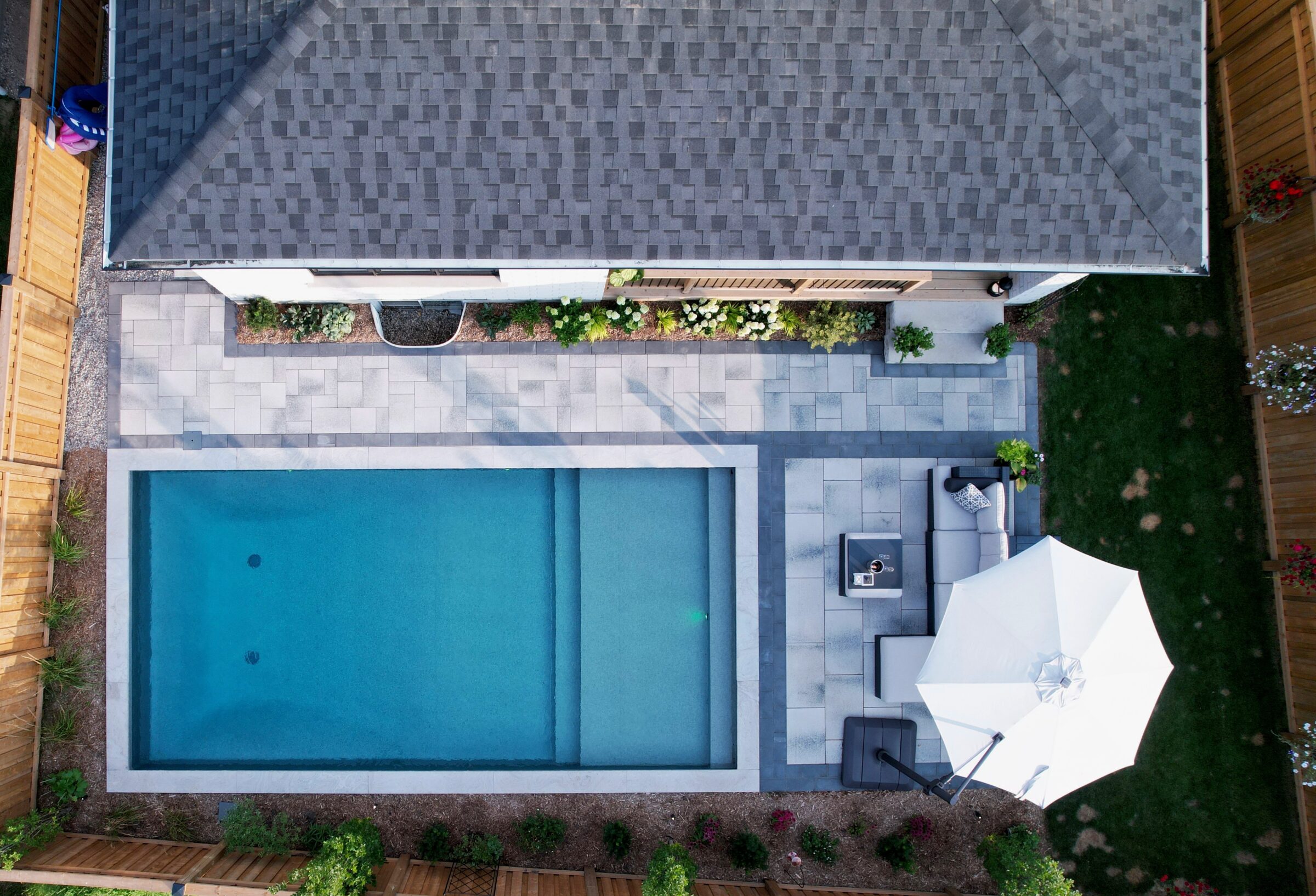 Aerial view of a modern backyard featuring a rectangular swimming pool, patio furniture, umbrella, and neatly landscaped garden surrounded by wooden fencing.