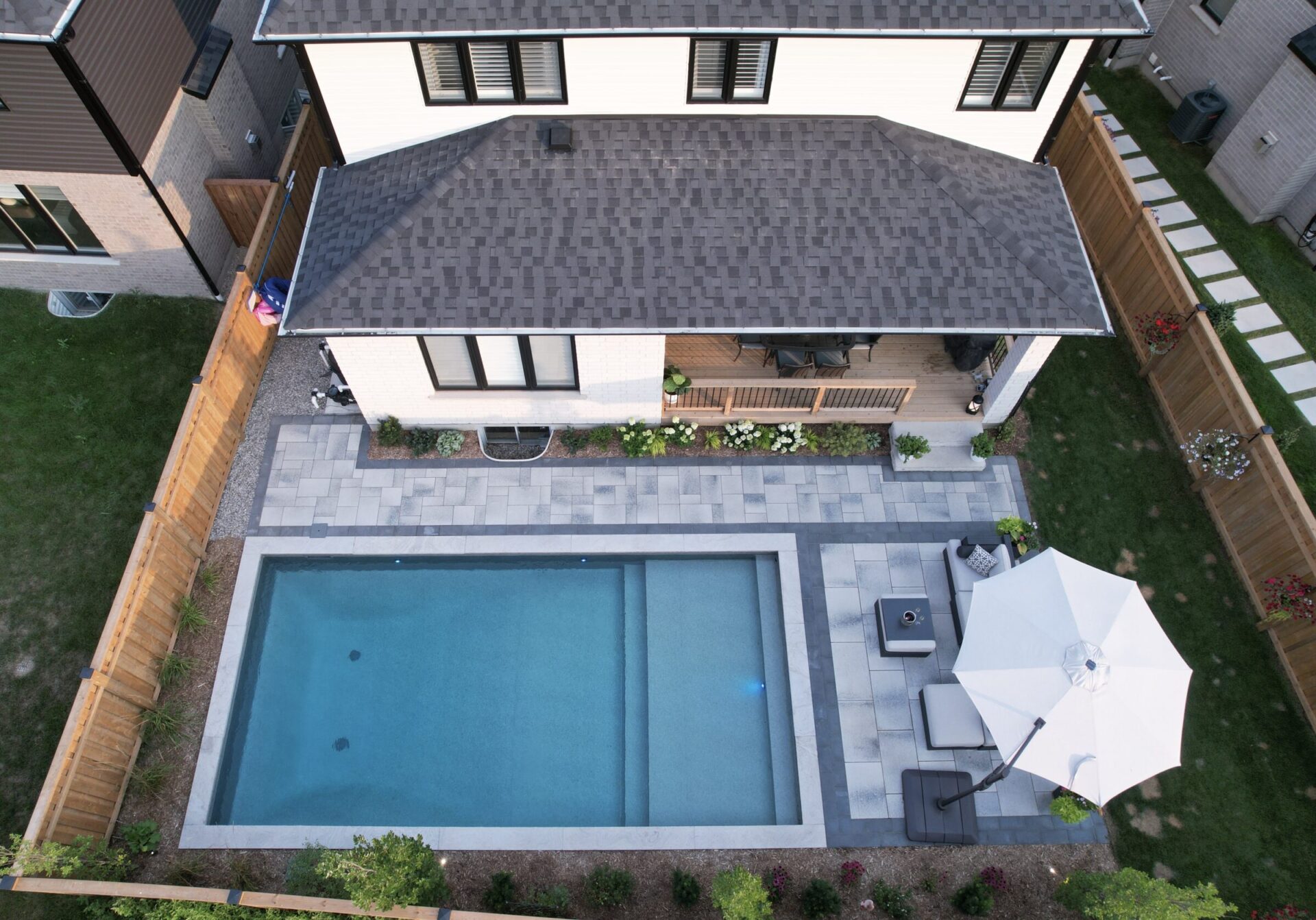Modern backyard with rectangular pool, patio furniture, and white umbrella. Surrounded by wooden fence, the area has plants and a deck.