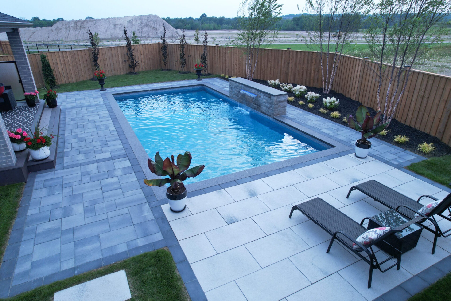 A modern backyard features a rectangular swimming pool, surrounded by sleek paving, flowers, and lounge chairs, set against a wooden fence and grassy area.