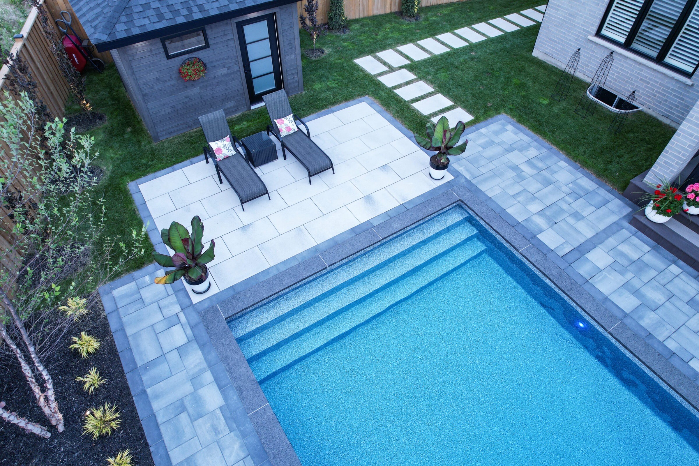 A backyard with a modern pool, lounge chairs, potted plants, and a wooden shed, surrounded by a small garden and patio.