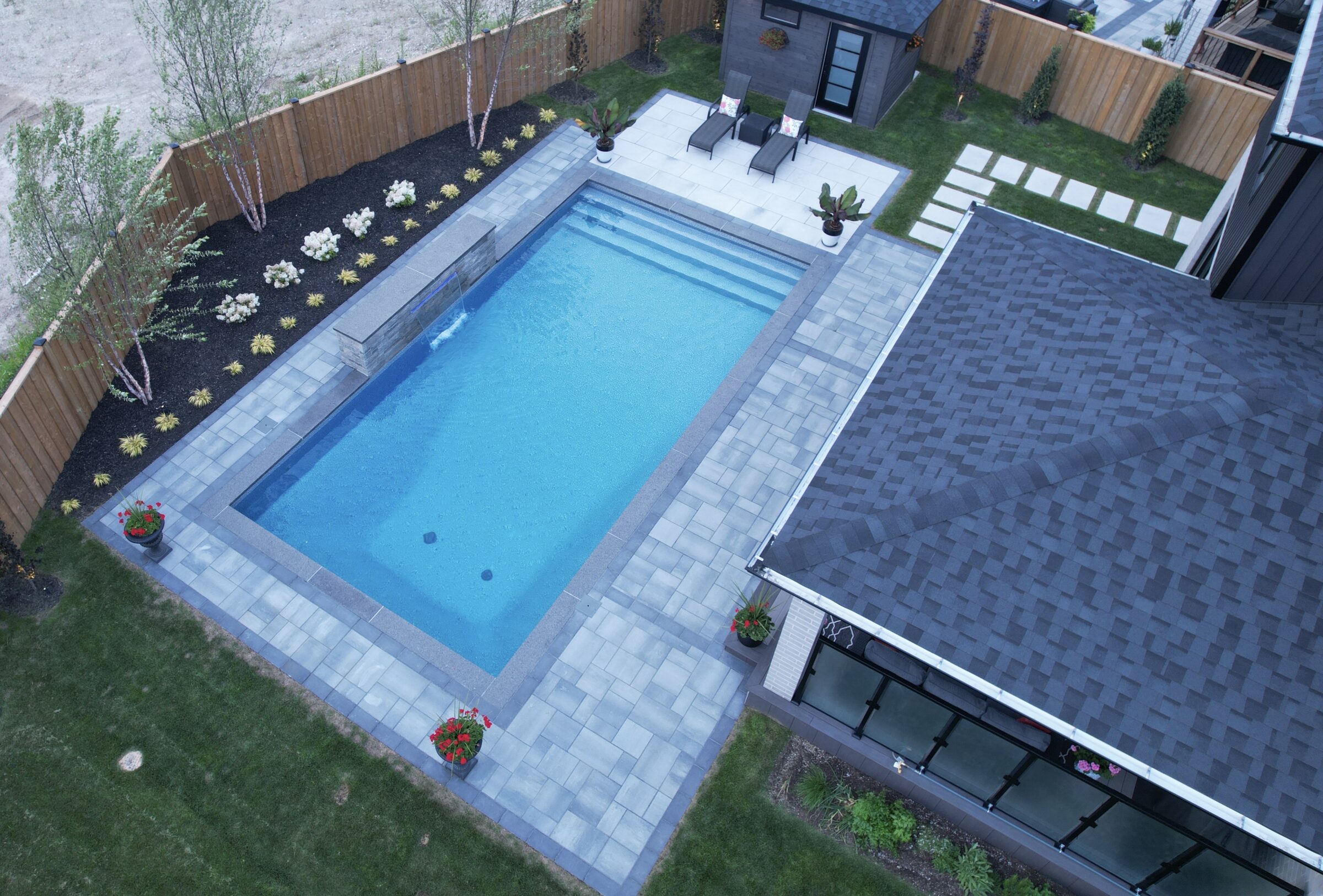A backyard scene with a rectangular swimming pool, patio furniture, landscaped garden, and adjacent building. No landmarks or historical buildings present.