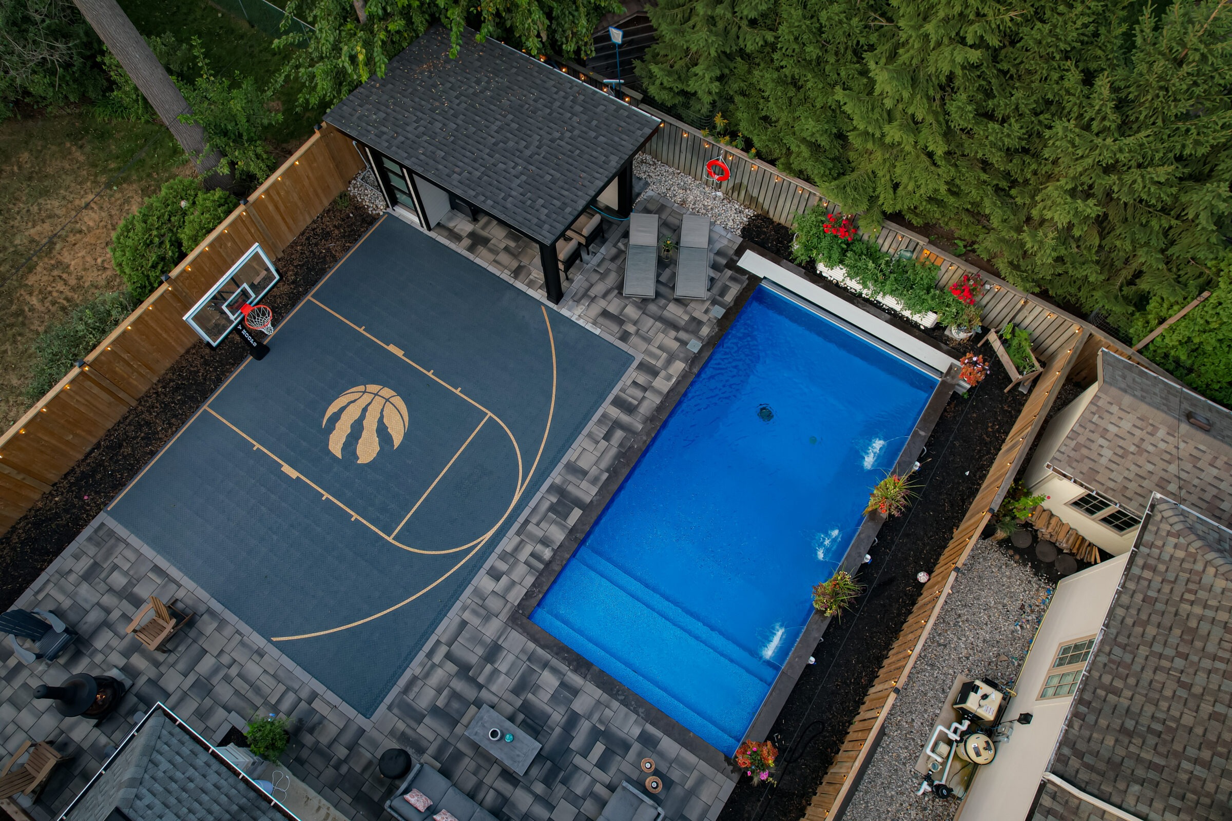 Stylish backyard features a basketball court, swimming pool, patio chairs, and a small building, surrounded by greenery and a wooden fence.