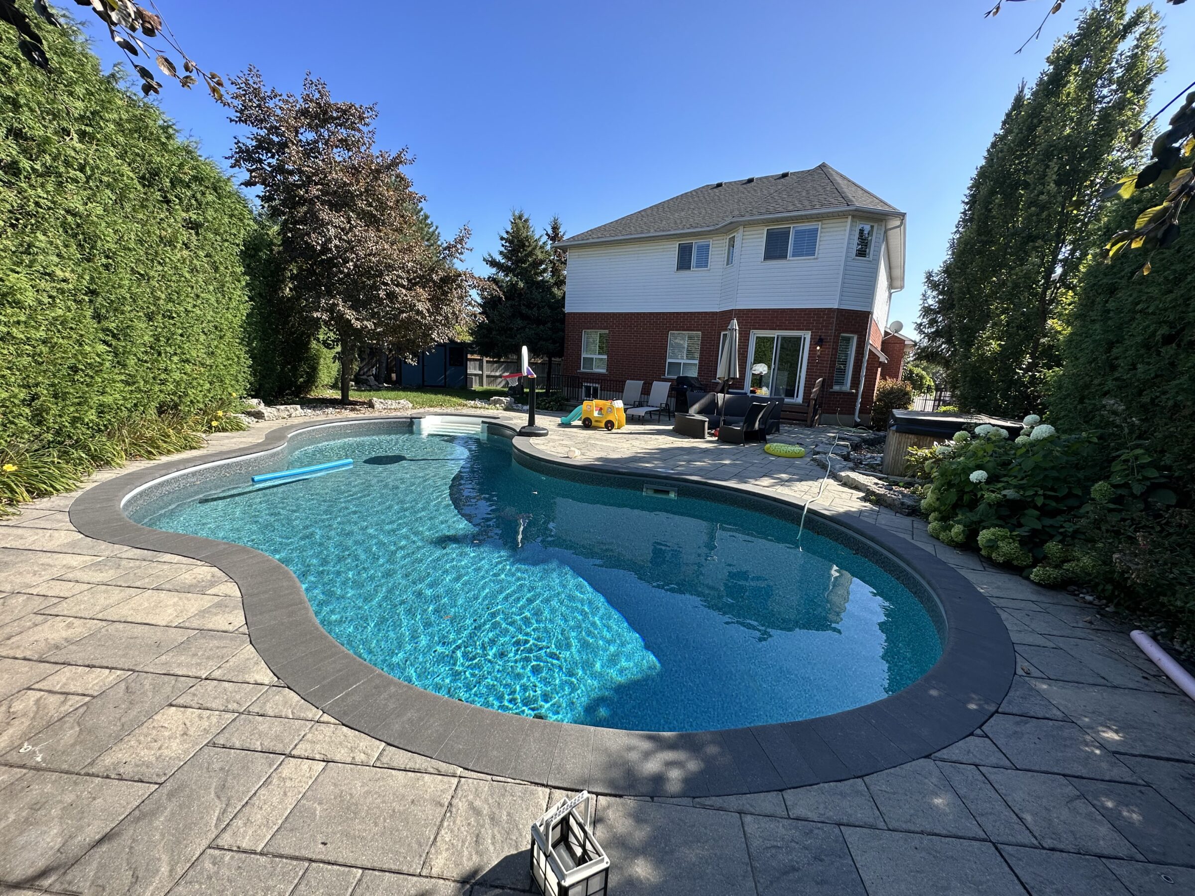 A curved swimming pool with clear water, surrounded by patio furniture and greenery, adjacent to a two-story brick house under a clear sky.