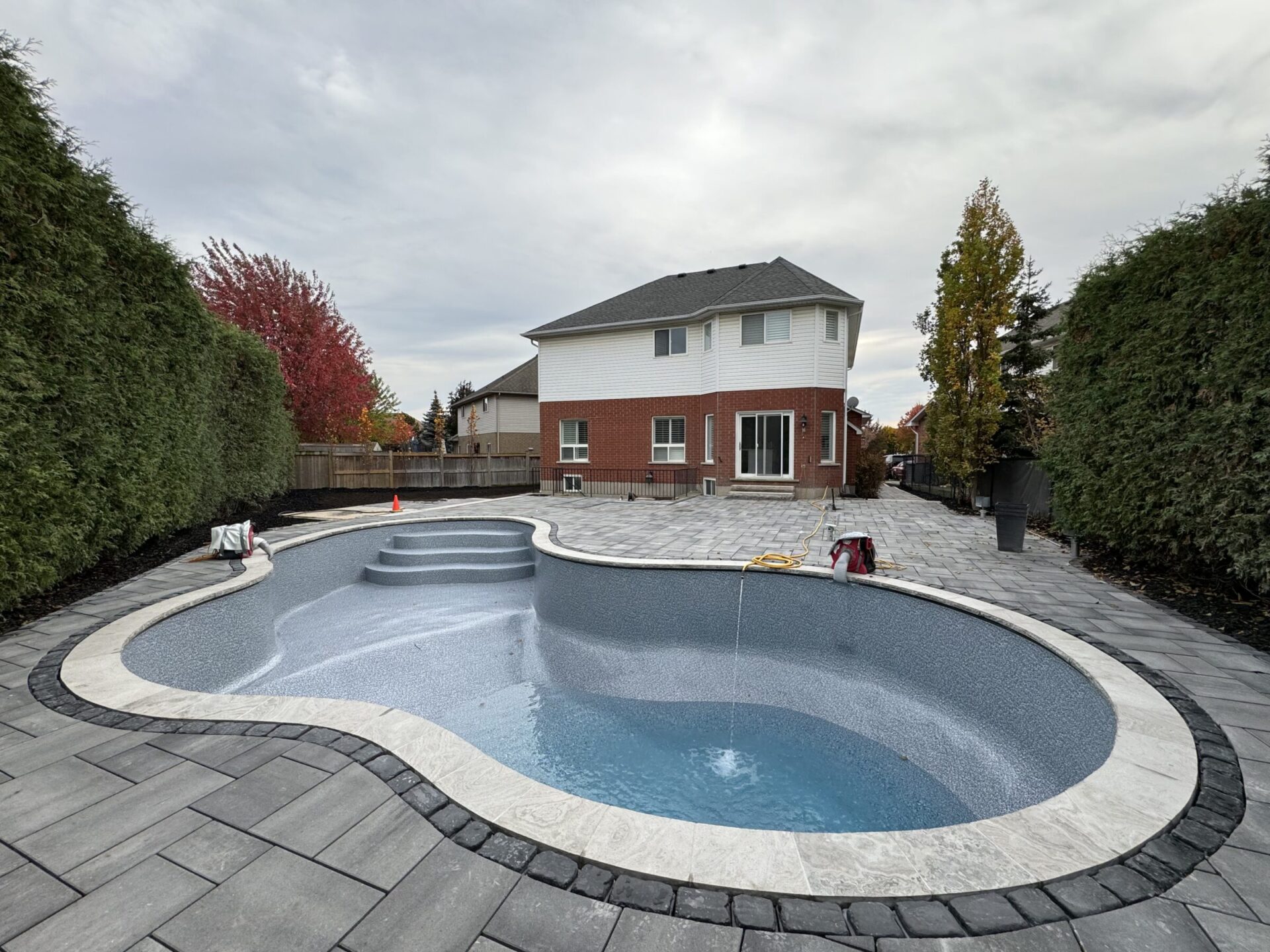 A backyard with an empty in-ground pool surrounded by stone tiles, framed by tall hedges and a brick house in the background.