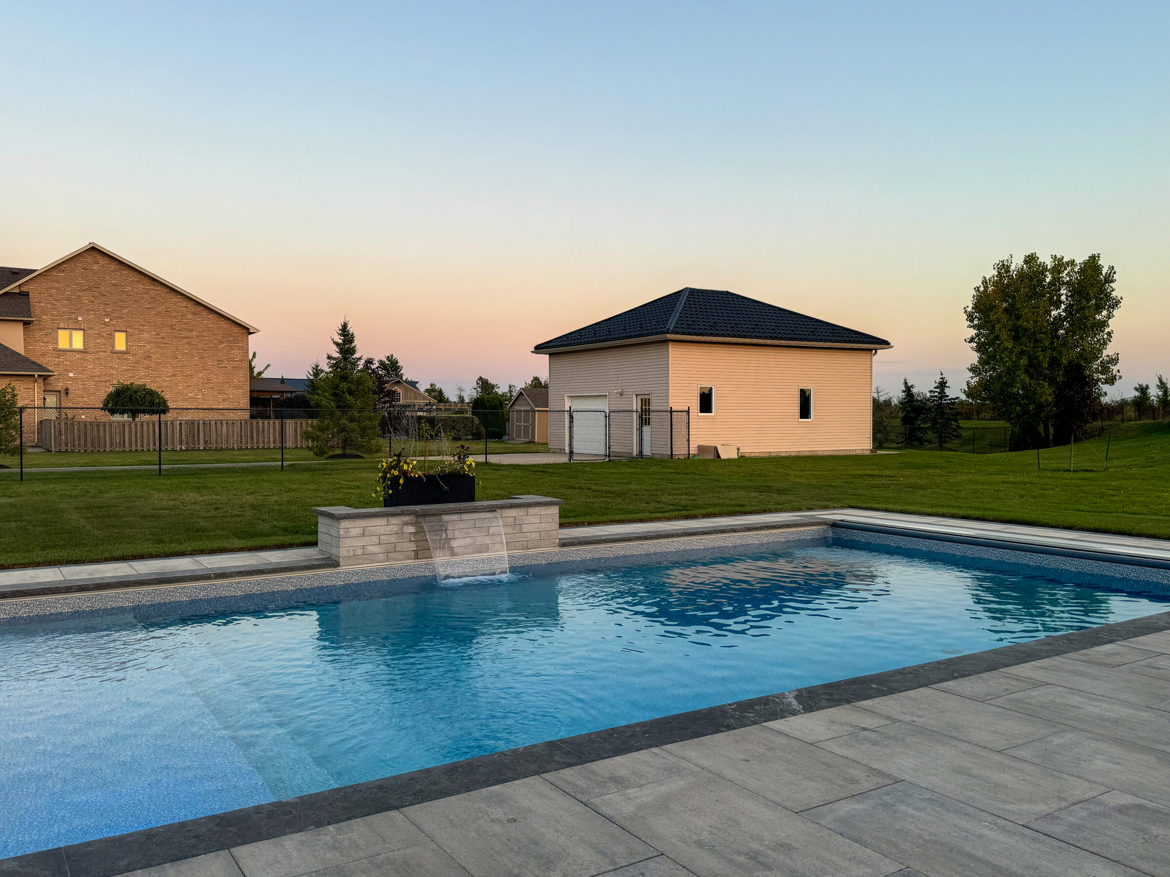 A serene residential backyard featuring a swimming pool, manicured lawn, two houses, and open sky during twilight, creating a peaceful ambiance.