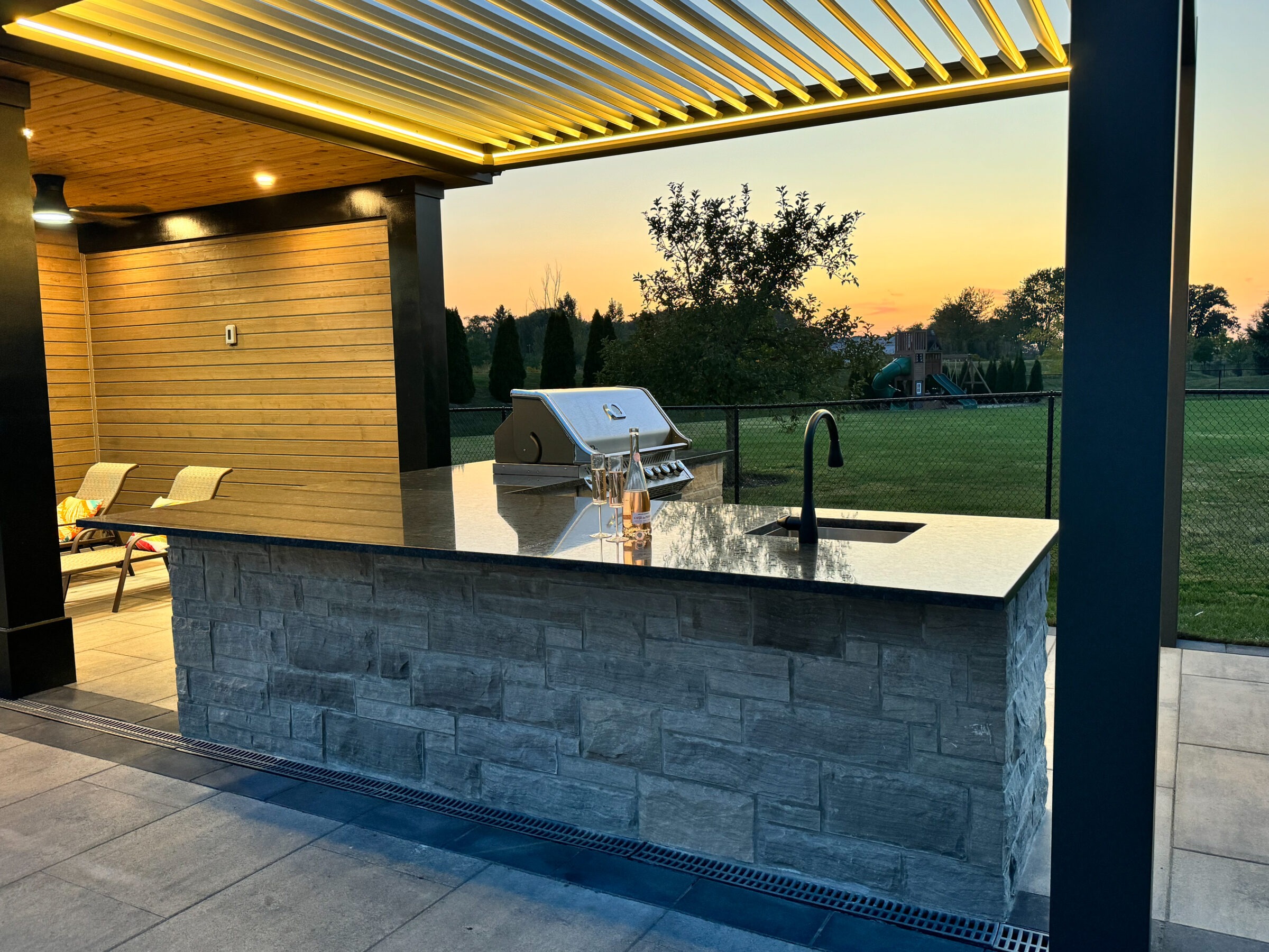 Modern outdoor kitchen with stone counter, grill, and sink under a pergola, overlooking a playground and grassy field at sunset. No people present.