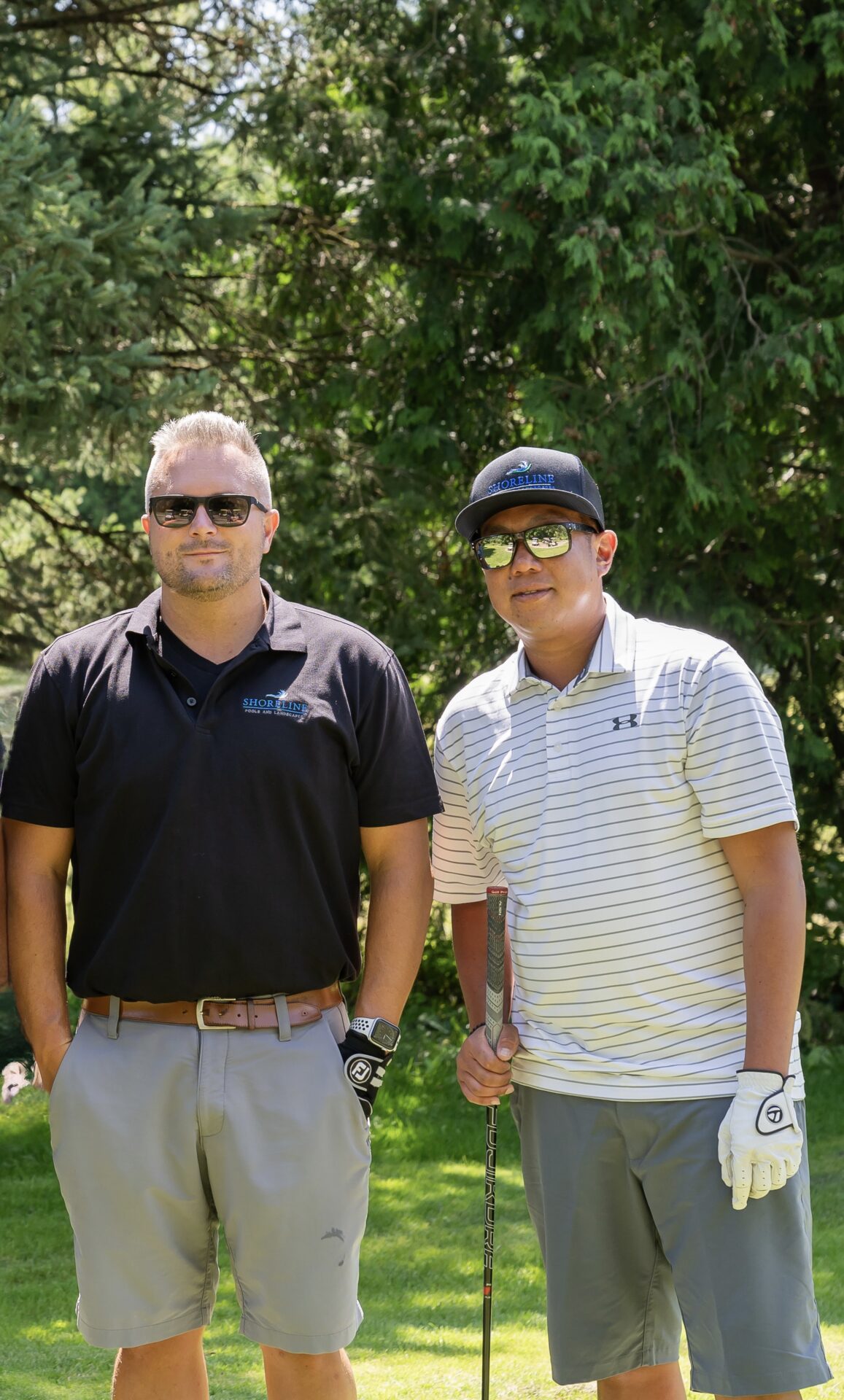 Two people wearing sunglasses and golf attire pose outdoors with trees in the background, holding a golf club and smiling.