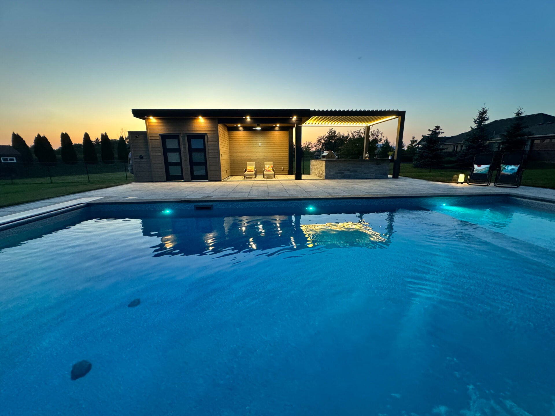 A serene backyard scene depicts a modern pool house with lounge chairs and a glowing swimming pool at sunset, surrounded by trees.