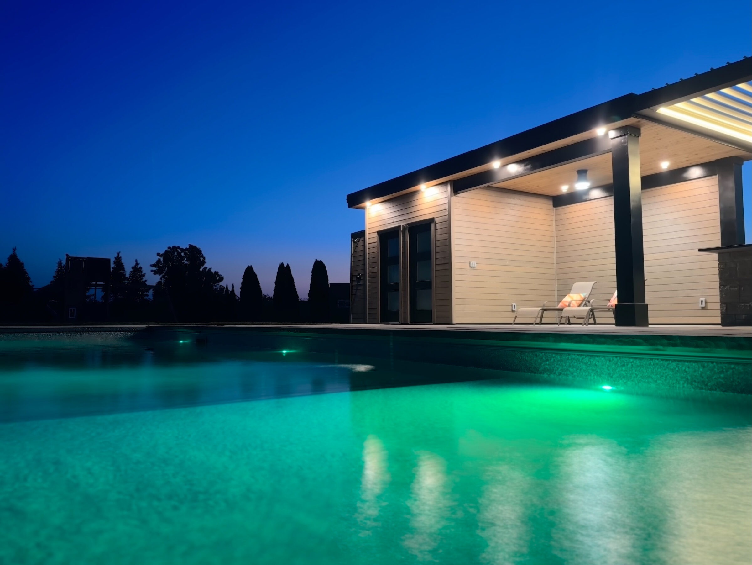 Modern poolside evening scene; illuminated water, sleek wooden structure, lounge chairs; serene ambiance under deep blue sky, surrounded by silhouetted trees.