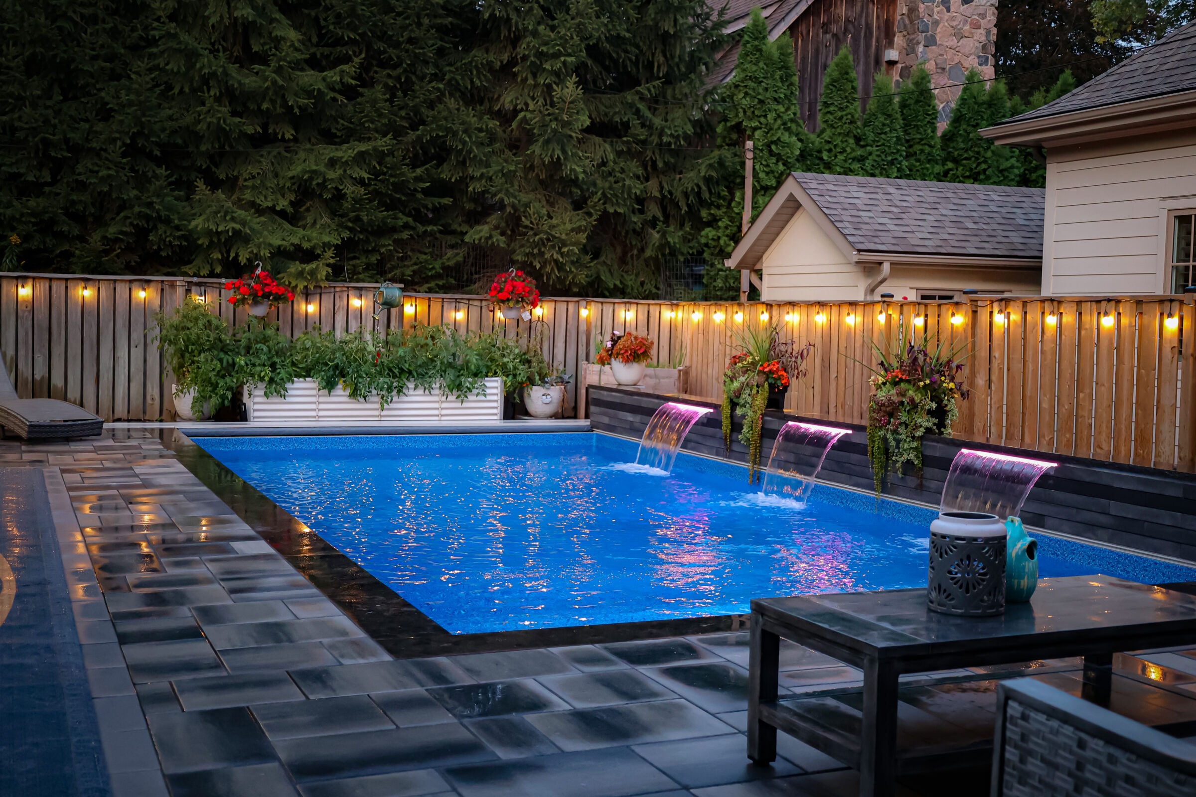 A serene backyard pool features cascading water, surrounded by plants and warm lights. Patio furniture sits nearby, enhancing the cozy ambiance.