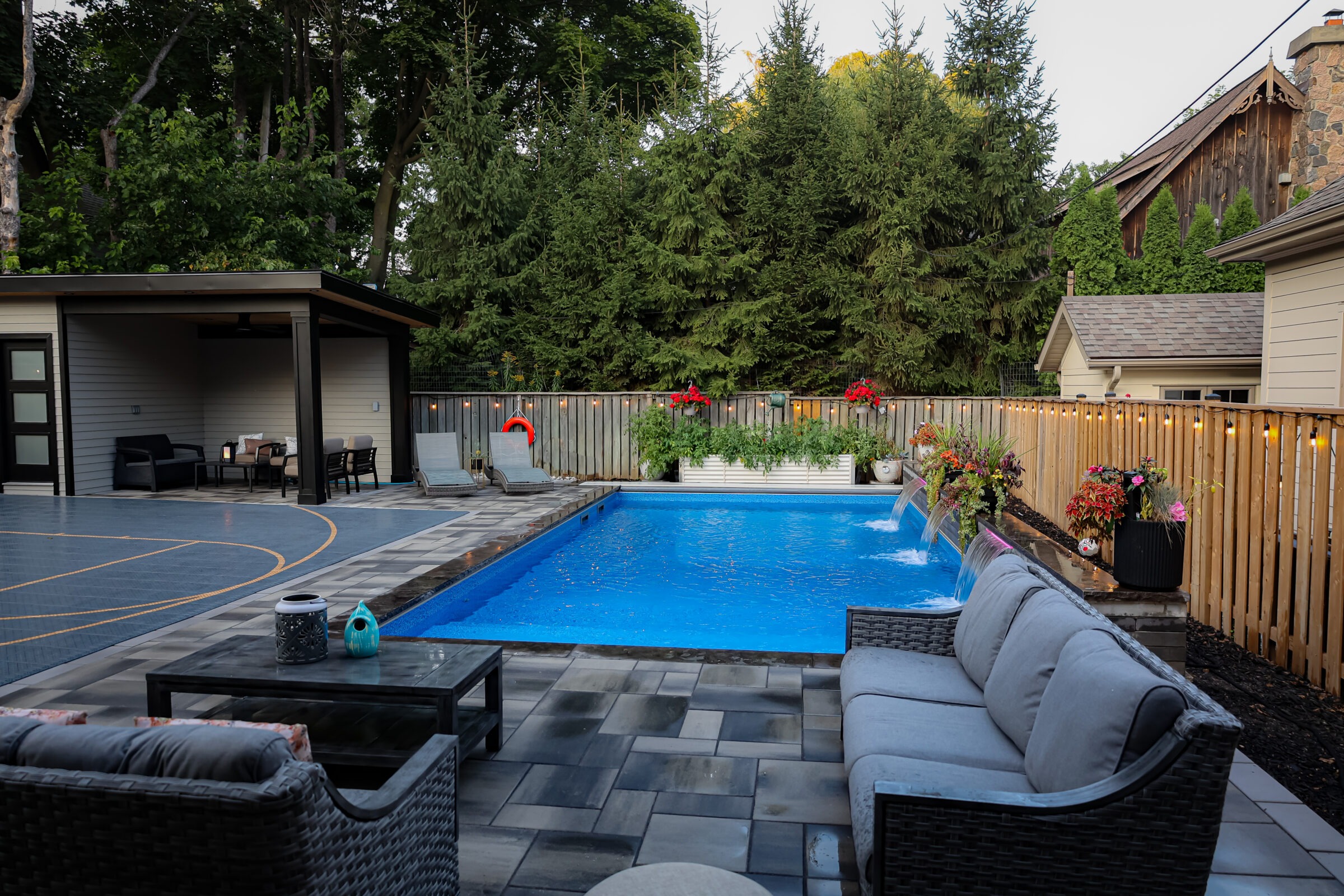 Cozy backyard with a blue pool, wicker furniture, a patio area, and lush greenery. A basketball court is visible beside a seating area.