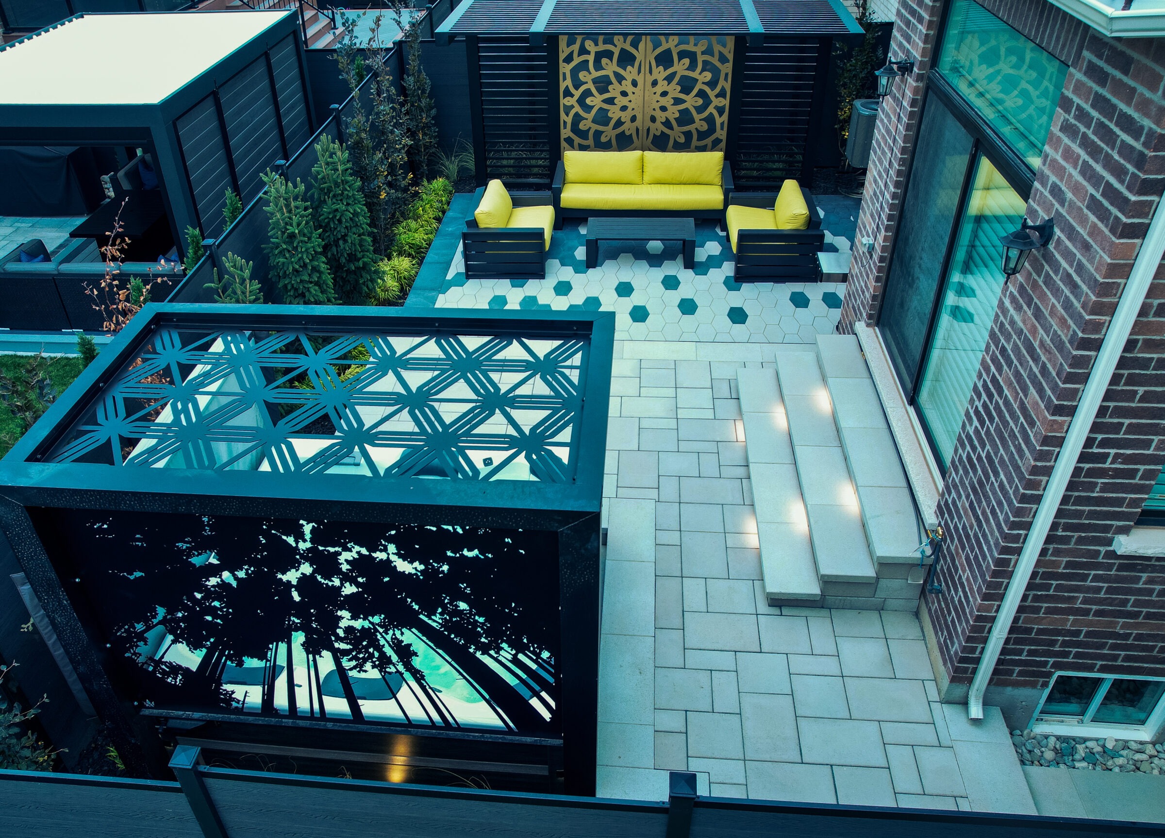 Modern outdoor patio with geometric designs, yellow seating, decorative panels, and neatly arranged plants. Steps lead to a glass door on brick wall.